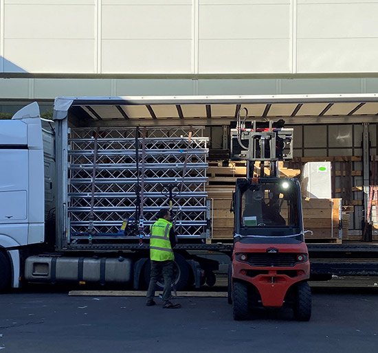 Röd lasttruck framför lastbil med trälådor som innehåller mässartiklar